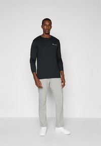 Long-sleeve black t-shirt with a small white logo, paired with light gray sweatpants and white sneakers, standing against a plain background.