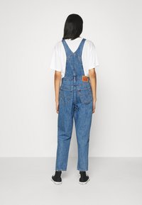 Denimoverall med justerbara remmar, stora bakfickor och en läderlogotyp, i kombination med en vit skjorta och svarta sneakers.