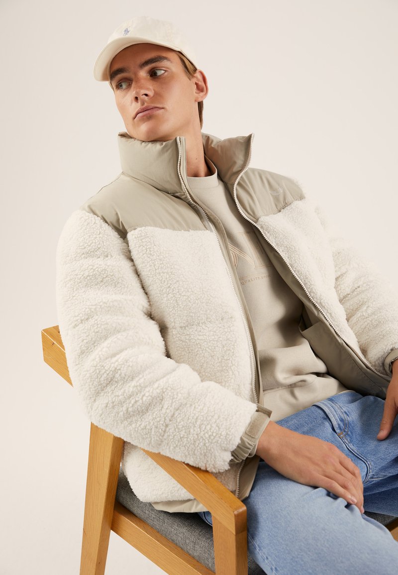 Roomkleurige schapenvachtjas met een lichtbeige nylon bovenkant, hoge kraag en ritssluiting. Draag het over een beige sweatshirt en lichtblauwe spijkerbroek.