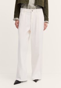 Eine Person trägt weiße, weit geschnittene Hosen, kombiniert mit einer gestreiften Bluse und einer olivgrünen, cropped Jacke, und steht vor einem neutralen Hintergrund.