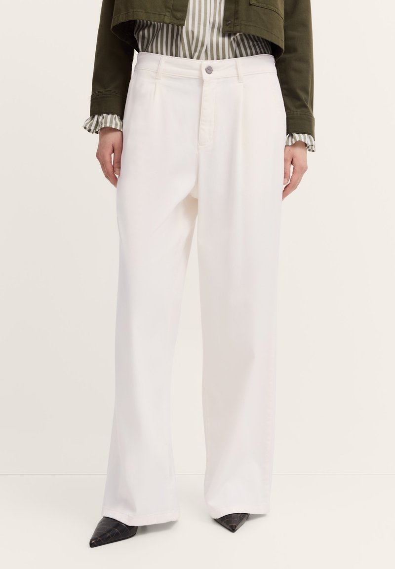Eine Person trägt weiße, weit geschnittene Hosen, kombiniert mit einer gestreiften Bluse und einer olivgrünen, cropped Jacke, und steht vor einem neutralen Hintergrund.