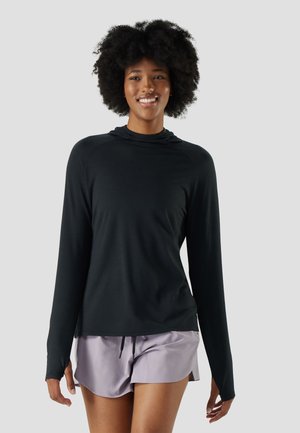 Smiling woman with curly hair wearing a black long-sleeve hoodie and lilac running shorts against a plain background.