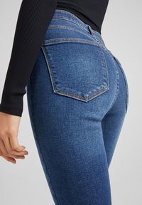 Blå denimjeans med hög midja, figursydd passform och två bakfickor. Tyget har en lätt textur och en medelvättad look.