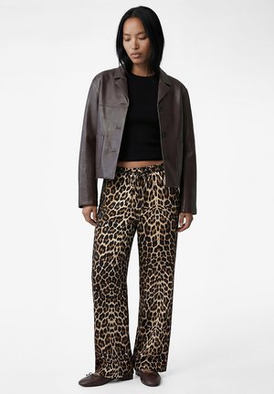 Femme portant une veste en cuir marron foncé, un haut court noir, un pantalon ample à motif léopard, et des ballerines marron foncé, debout devant un fond blanc.