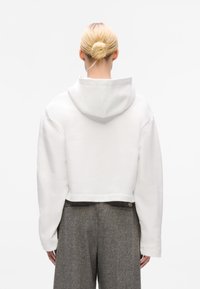 Cropped, white hoodie with a soft texture and a hood. Features dropped shoulders and a straight hem. Worn over gray high-waisted trousers.