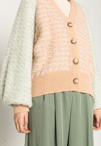 Beżowy i miętowozielony sweter na guziki z teksturowanym wzorem, drewnianymi guzikami i prążkowymi mankietami. Noszony na zielonej bluzce.