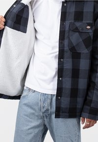 Svart och marineblå rutiga skjorta med fleecefoder, med tryckknappar, en bröstficka, bärs över en vit t-shirt och ljusblå jeans.