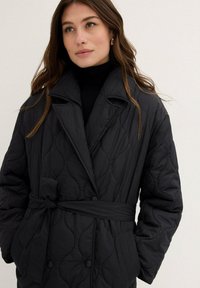 Cappotto imbottito nero con cintura, colletto oversize, modello a trama testurizzata e tasche laterali. Presenta grandi bottoni e un design elegante e moderno.