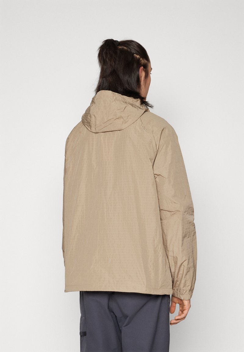 Chaqueta ligera beige con capucha, de tela texturizada, con puños elásticos y corte holgado, vista desde la parte posterior.