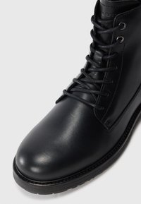 Black leather lace-up boot with a rounded toe, smooth texture, sturdy sole, and silver eyelets. Top features a logo detail on the tongue.