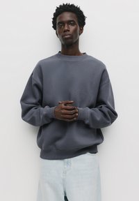 Mørkegrå oversized sweatshirt med rund halsudskæring og ribbede manchetter, parret med lyseblå denimjeans. Glat tekstur, minimalistisk design.