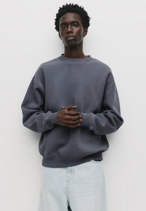 Dark gray oversized sweatshirt with a round neckline and ribbed cuffs, paired with light blue denim jeans. Smooth texture, minimal design.