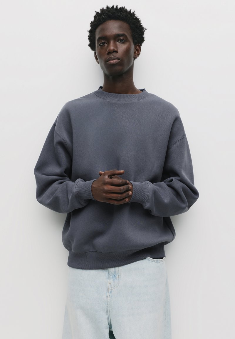 Mørkegrå oversized sweatshirt med rund halsudskæring og ribbede manchetter, parret med lyseblå denimjeans. Glat tekstur, minimalistisk design.