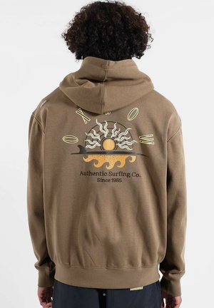 Sweat à capuche en polaire marron avec un graphisme soleil et vague au dos, accompagné du texte "Authentic Surfing Co. Depuis 1985". Matière douce et coupe décontractée.
