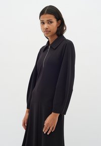 Čierny zip-up top s golierom a dlhými rukávmi, s hladkou textúrou a jemným objemom v rukávoch.