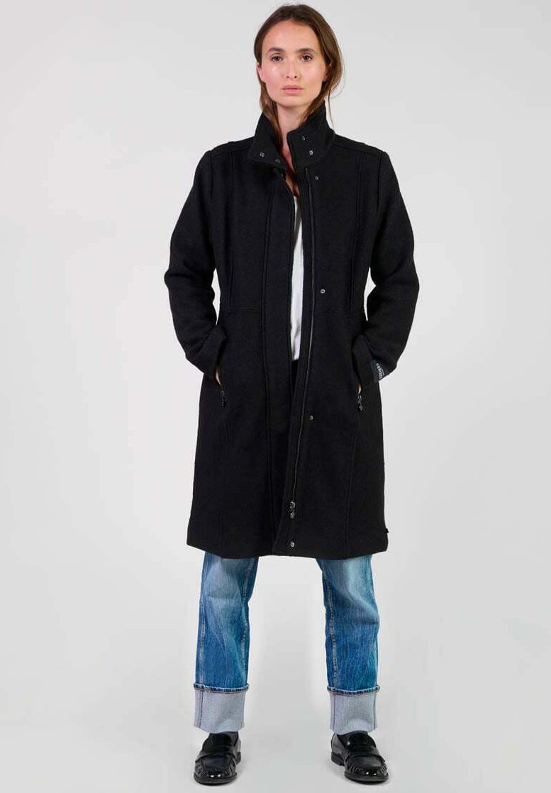 Manteau long noir avec un col haut, fermeture éclair devant, poches latérales et tissu texturé. Associé à un jean bleu et des chaussures noires.