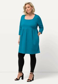 Tyrkysové šaty s čtvercovým výstřihem a tříčtvrtečními rukávy, s nařaseným pasem a bočními kapsami, v kombinaci s černými legínami a podpatky.