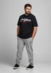 Černé bavlněné tričko s bílým a červeným logem, doplněné světle šedými tepláky s logem na nášivce. Model má na sobě černé boty.