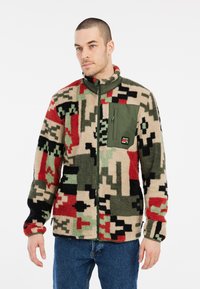 Fleece jakna u bež, crnoj, crvenoj i zelenoj kamuflažnoj uzorku; sadrži zeleni džep na zatvarač na prsima i visoki ovratnik.