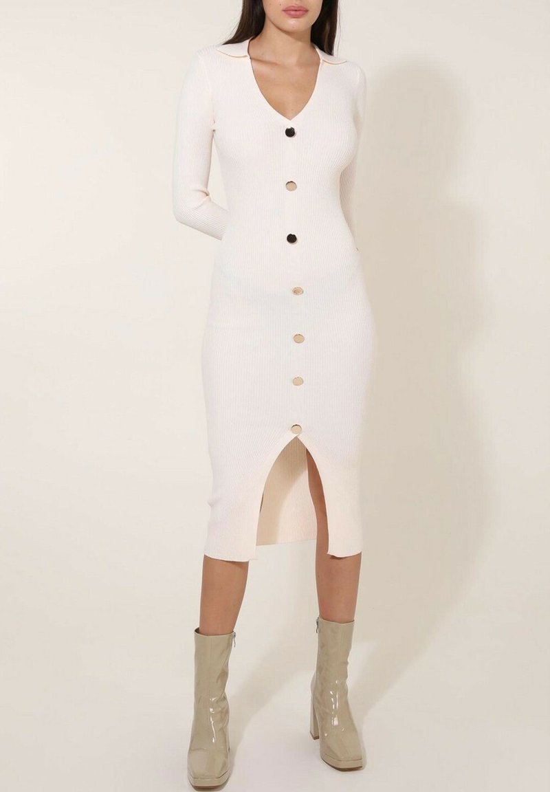 Robe en maille côtelée ajustée en blanc cassé avec des manches longues, un décolleté en V, un détail de bouton sur le devant et une fente cuisse ; associée à des bottines beiges brillantes.