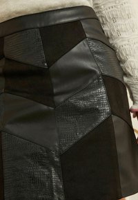 Melna svārku dizains ar ģeometrisku laika patchwork, kas prezentē dažādas tekstūras: gluda āda un reljefs kroko prints; pieguļošs jostasvieta.