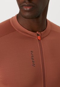 Maglietta ciclismo a maniche corte color ruggine. Presenta un tessuto testurizzato, una zip parziale con una linguetta arancione e il marchio "Isadore" in nero.