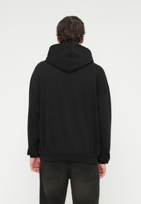 Μαύρο φούτερ με μεγάλο κουκούλι, χαλαρές ραφές στους ώμους και άνετη εφαρμογή, με μονόχρωμο σχέδιο και απαλή υφή. Η πίσω όψη δείχνει απλότητα.