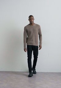 Bruine gebreide trui met een ronde halslijn, gedragen over zwarte slim-fit jeans en zwarte schoenen, weergegeven tegen een lichtgrijze achtergrond.
