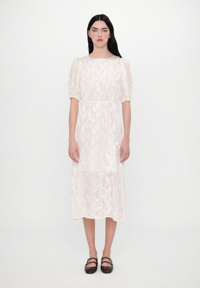 Cremfarbendes Midi-Kleid mit transparenter Blumentextur, Puffärmeln, rundem Ausschnitt und tailliertem Gürtel. Kombiniert mit dunkelbraunen Schuhen.