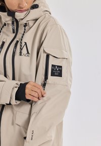 Beige Außenjacke mit hohem Kragen, schwarzen Akzenten, mehreren Reißverschlusstaschen und markenbezogenen Patches an den Ärmeln. Wasserdichtes Material.