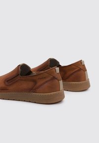 Zapatos marrones de cuero sin cordones con textura perforada y paneles laterales elásticos. Suelas de goma color caqui y un sutil logotipo en el talón.