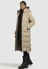 Lungo cappotto imbottito beige con cappuccio, caratterizzato da dettagli neri, una cerniera frontale e tasche laterali. Abbinato a stivali da combattimento neri con lacci.