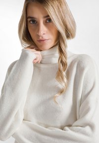 Pulover alb cu guler înalt, realizat dintr-un material texturat și moale. Are mâneci lungi și o croială lejeră. Există un model subtil în țesătură.