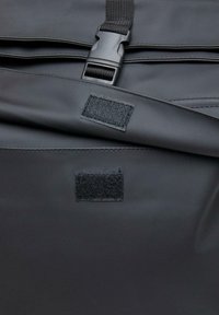 Mustapintainen vedenpitävä laukku, jossa on matta pinta, säädettävät hihnat ja kaksi tarranauhapistettä kiinnityksiä tai sulkemista varten.