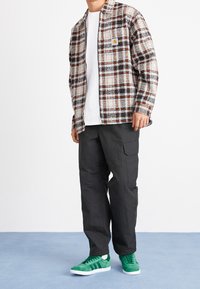 Geruit flanellen overhemd in beige, zwart en oranje; wit T-shirt eronder; cargobroek in zwart; groene suède sneakers met witte zolen.