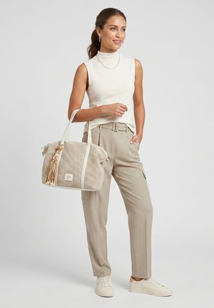 Kvinde i hvid ærmeløs top og beige bukser med bælte står og holder en beige flettet håndtaske med kvast, iført hvide sneakers.