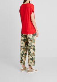 T-shirt rossa a maniche corte abbinata a pantaloni cargo larghi in camouflage verde e beige, completata da sneakers color marrone chiaro.