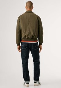 Chaqueta bomber verde oliva con puños y dobladillo acanalados, llevada sobre una camisa roja y unos vaqueros azul oscuro, que muestra un diseño casual pero ajustado.