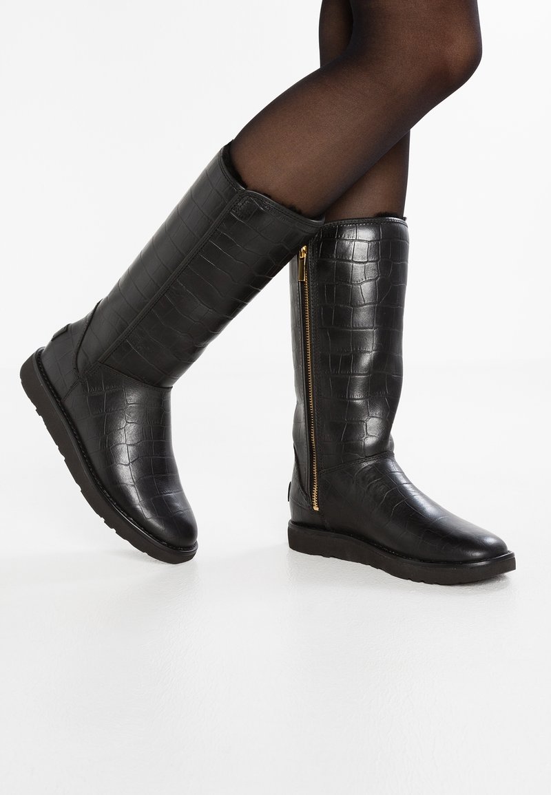 Femme portant des bottes noires montantes avec texture crocodile et fermeture éclair latérale dorée, associées à des collants noirs translucides.