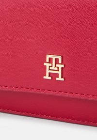 Rød læderpung med en struktureret overflade, prydet med en guldfarvet logoaccent og synlige sømdetaljer. Kompakt rektangulær form.