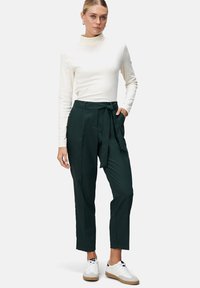 Witte coltrui en donkergroene broek met riem en hoge taille. Het model draagt witte sneakers. Gladde stof, nauwsluitende pasvorm en zijzakken.