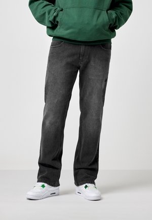 Personne portant un jean noir coupe décontractée, un sweat à capuche vert avec les mains dans les poches, et des baskets blanches avec des détails verts, debout sur un fond clair.