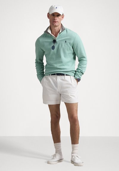 Grön långärmad tröja med krage, bärs över en vit polo; tillsammans med vita shorts, vita sneakers och en vit keps. Lätt tygkvalitet.