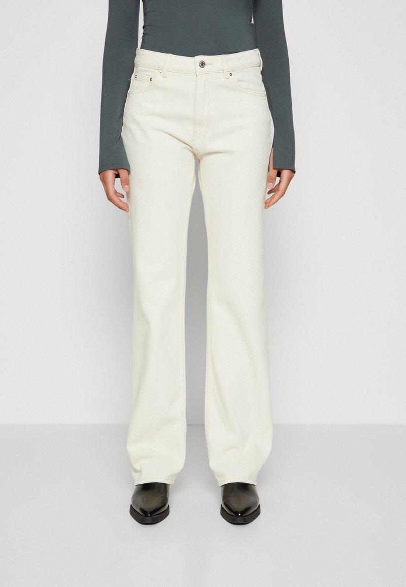 Jeans blancos acampanados de mezclilla, con cierre frontal de botones, trabillas para cinturón y bolsillos delanteros, combinados con una camiseta ajustada de manga larga.