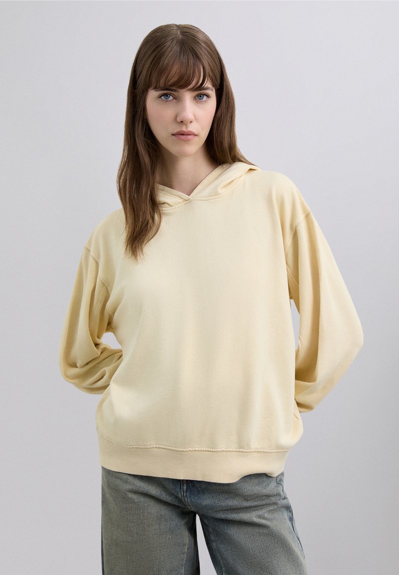 Gelber Kapuzenpullover aus weichem Stoff, mit einer Fronttasche, einer Kordelzugkapuze und übergroßen Ärmeln. Kombiniert mit hellblauen Jeans.