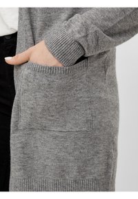 Hand with painted nails inside the front pocket of a grey knitted cardigan with ribbed cuffs and hem.