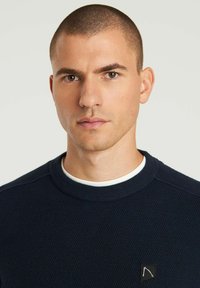 Marineblauer, strukturierter Pullover mit Rundhalsausschnitt, ausgestattet mit einem weißen Innenkragen und einem schwarzen Logopatch auf der Brust. Angenehmer Stoff mit figurbetontem Design.