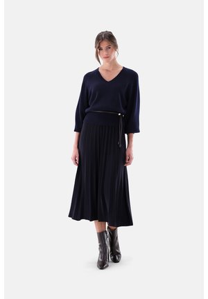 Marineblauer V-Ausschnitt-Pullover mit dreiviertellangen Ärmeln, kombiniert mit einem plissierten marineblauen Midirock, ergänzt durch schwarze Stiefeletten und einen dünnen Gürtel.