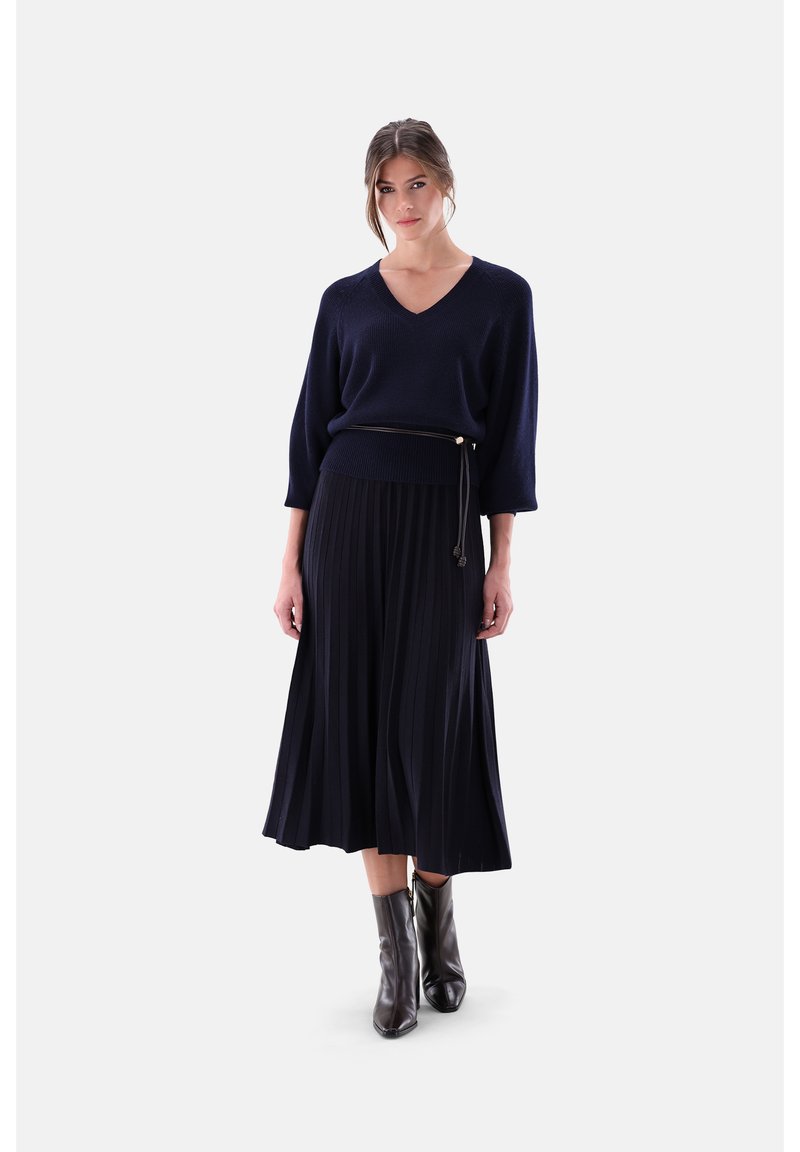 Marineblauer V-Ausschnitt-Pullover mit dreiviertellangen Ärmeln, kombiniert mit einem plissierten marineblauen Midirock, ergänzt durch schwarze Stiefeletten und einen dünnen Gürtel.