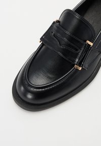Schwarze Lederloafer mit glatter Oberfläche, einem dekorativen Riemen mit goldfarbener Hardware und einem abgerundeten Zehenbereich.